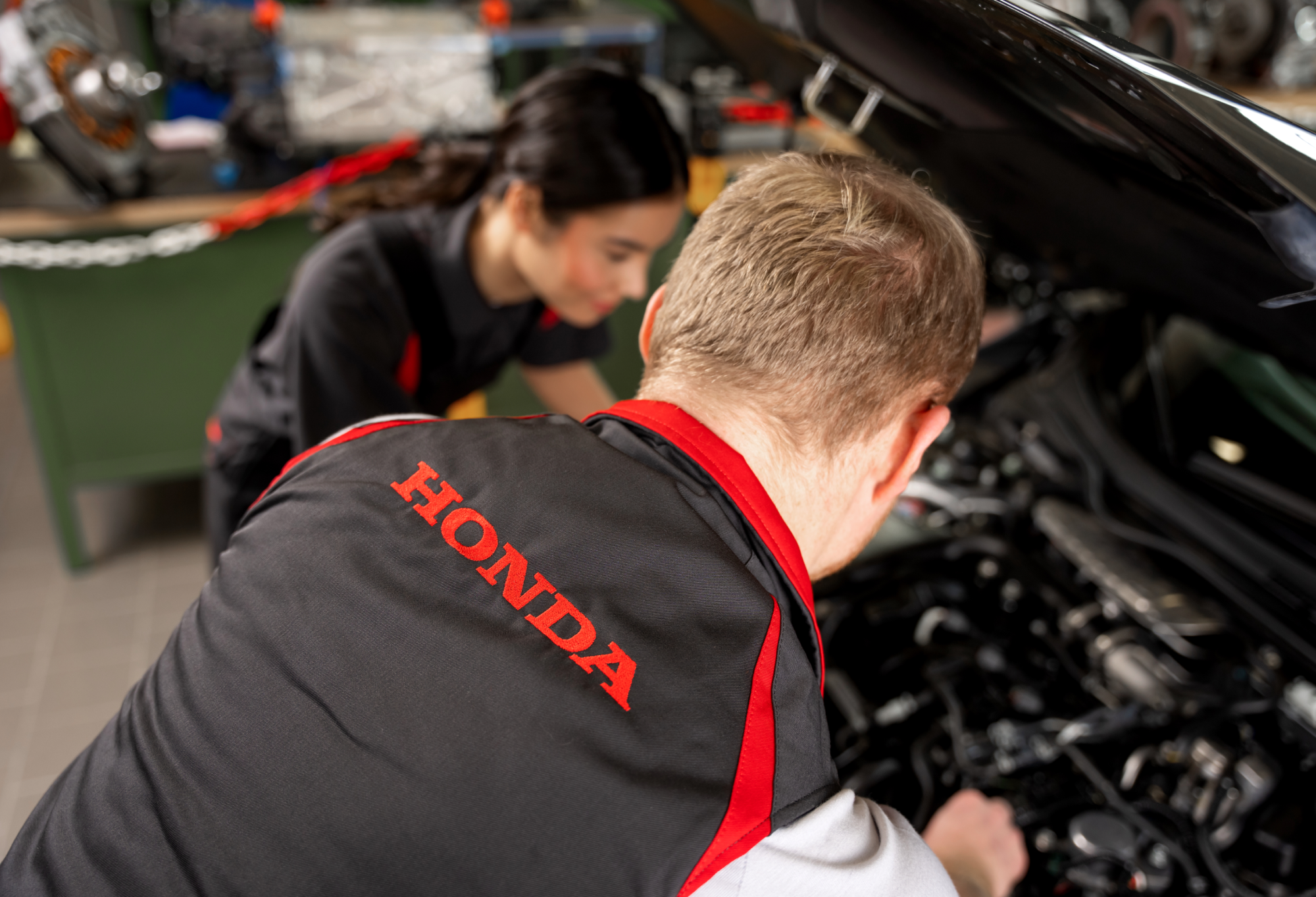 Two Honda mechanics working on a car engine. One mechanic, wearing a Honda-branded jacket, is leaning over the open hood, while another mechanic stands nearby observing.